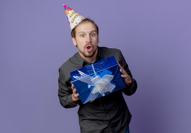 Surprised handsome man in birthday cap holds gift box isolated on purple wall