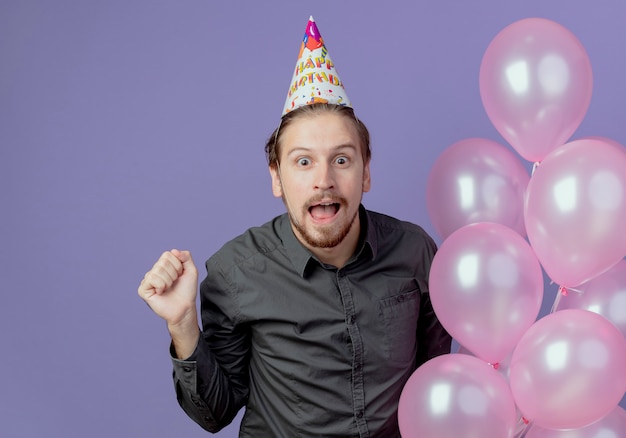 생일 모자에 놀란 잘 생긴 남자는 헬륨 풍선을 보유하고 보라색 벽에 고립 된 주먹을 유지