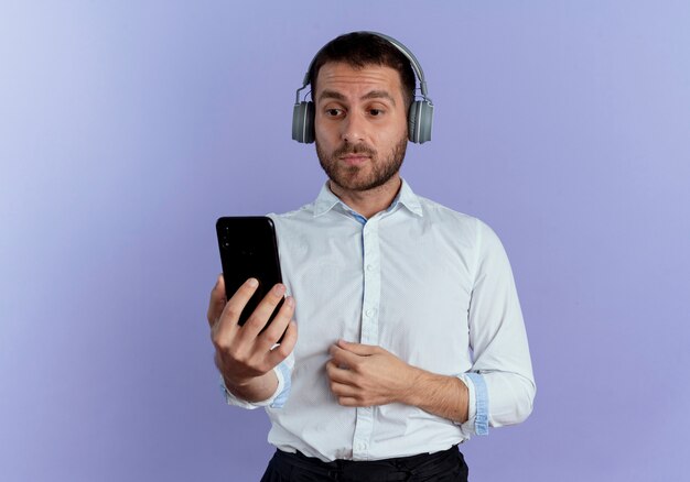Surprised handsome man on headphones holds and looks at phone isolated on purple wall