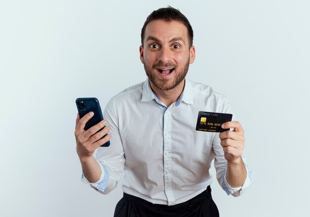 Surprised handsome man holds credit card and phone isolated on white wall