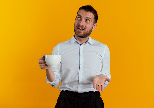 Surprised handsome man holds cup and looks up isolated on orange wall