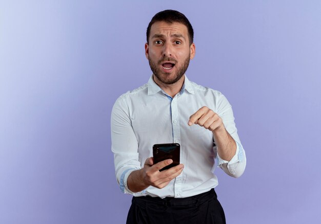 Surprised handsome man holds and points at phone isolated on purple wall