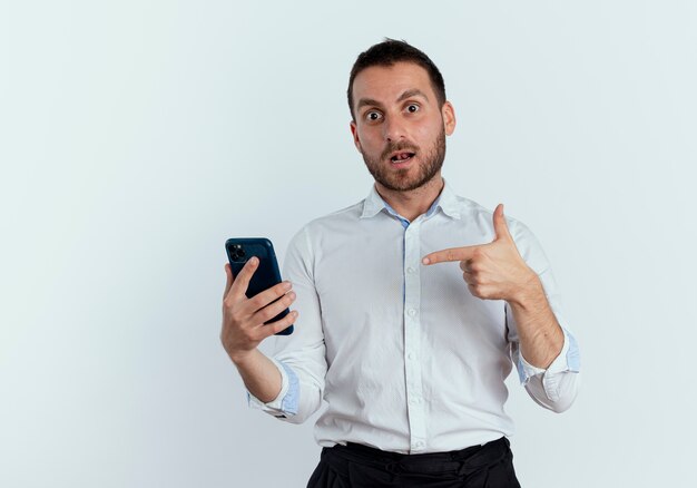 Surprised handsome man holds and points at phone isolated on white wall