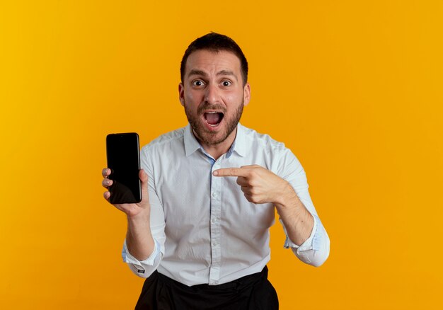 Surprised handsome man holds and points at phone looking isolated on orange wall