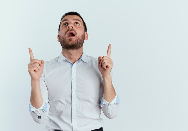 Surprised handsome man looks and points up with two hands isolated on white wall