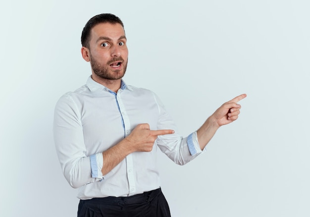 Surprised handsome man points at side with two hands looking isolated on white wall