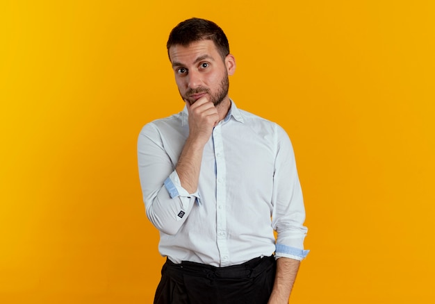 Surprised handsome man puts hand on chin isolated on orange wall