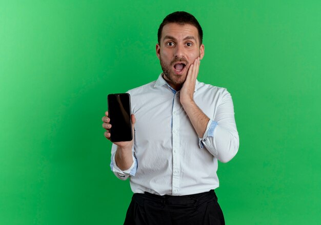 Surprised handsome man puts hand on face and holds phone isolated on green wall