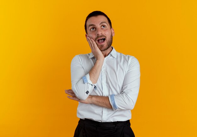 Surprised handsome man puts hand on face looking up isolated on orange wall