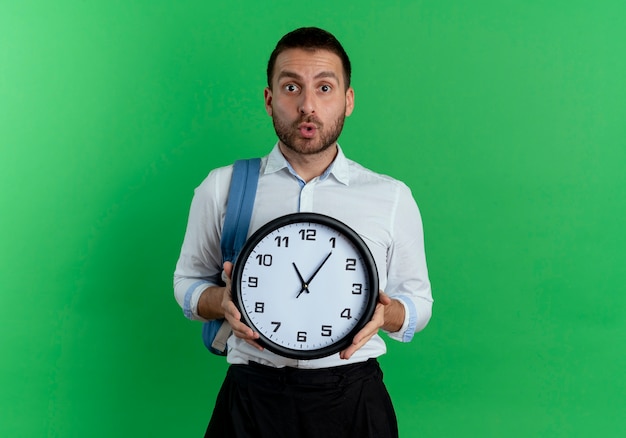 Surprised handsome man wearing backpack holds clock isolated on green wall