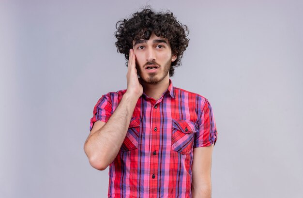 A surprised handsome man with curly hair in checked shirt holding hand on face 