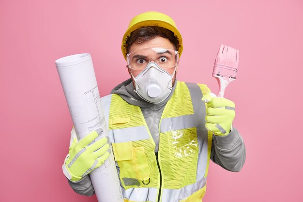 Surprised handyman holds blueprint and paint brush works on construction project plan wears protective helmet mask and glasses uniform 