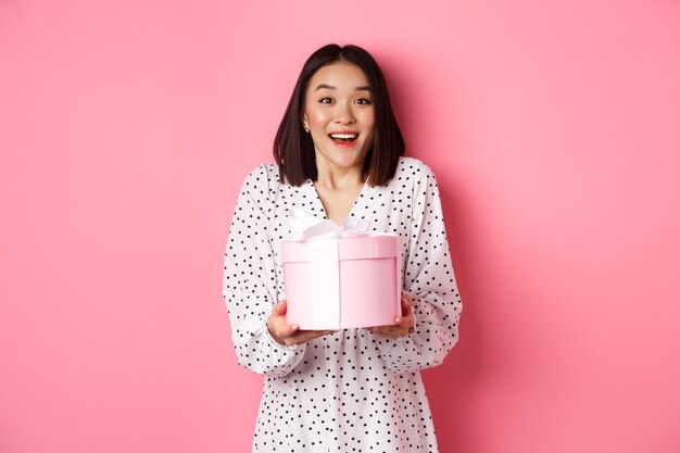 Surprised and happy asian woman receiving cute box with gift standing amazed over pink background