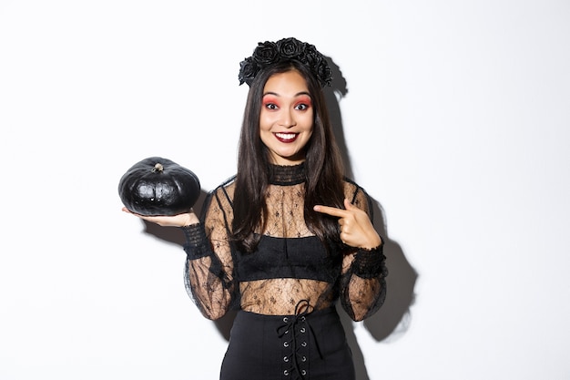 Free photo surprised happy asian woman in witch costume pointing finger at black pumpkin