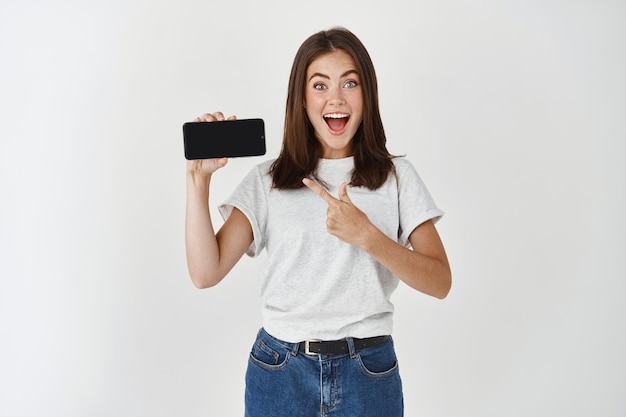 Удивленная счастливая брюнетка женщина в футболке показывает пустой экран смартфона и указывает на него, глядя в камеру с открытым ртом над белой стеной.