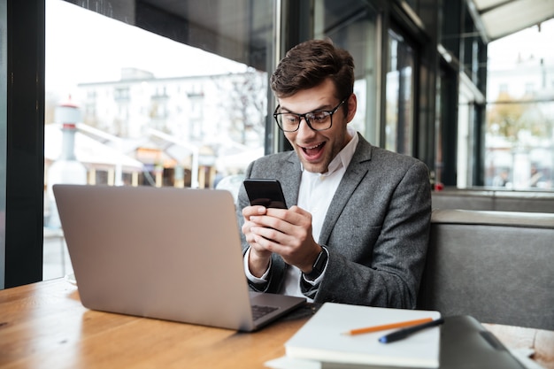 Surprised happy businessman in eyeglasses sitting by the table in cafe with laptop computer and using smartphone