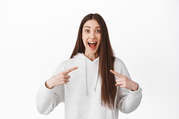 Surprised and happy candid girl pointing at herself raising eyebrows and gasping amazed being chosen as winner prize winning or achieve goal recieve good news standing over white background