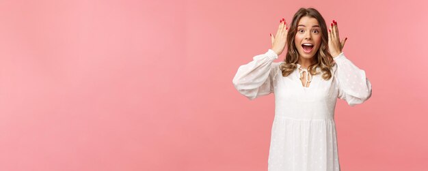 Surprised happy excited bday girl in white dress open her eyes to see incredible awesome gift from friends at party standing over pink background look astonished and cheerful