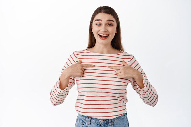 Surprised happy girl pointing at herself pleased to be chosen winning something hear great news about her standing over white background