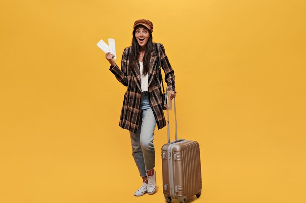 Surprised happy woman looks into camera and holds tickets Longhaired tanned girl in coat and jeans poses with suit bag on yellow background