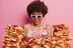 Free photo surprised happy woman points at herself and cannot believe her success, wears sunglasses and elegant clothing, poses over rosy wall in cafe, surrounded by sandwiches, going to have tasty lunch