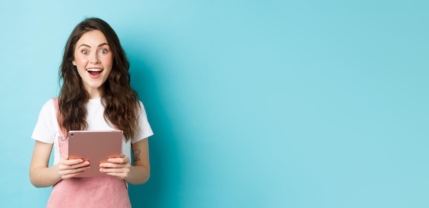 Surprised happy woman stare at camera amazed hear amazing news holding tablet in hands standing over blue background