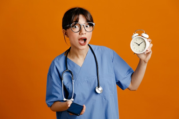 Free photo surprised holding alarm clock speaks on the phone young female doctor wearing uniform fith stethoscope isolated on orange background