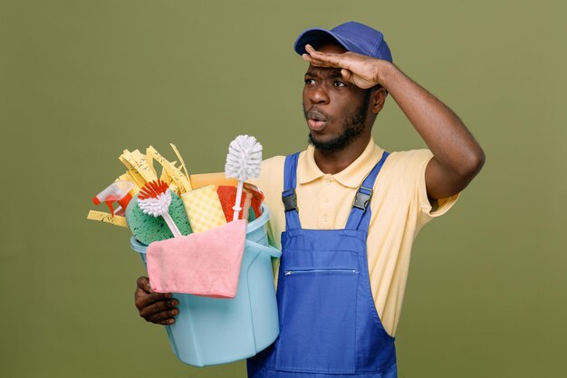 Surprised holding bucket of cleaning tools young africanamerican cleaner male in uniform with gloves isolated on green background