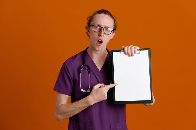 オレンジ色の背景に分離された聴診器で制服を着てクリップボードを保持している驚いた若い男性医師