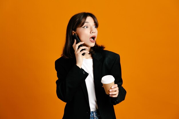 Surprised holding cup of coffee speaks on the phone young beautiful female wearing black jacket isolated on orange background