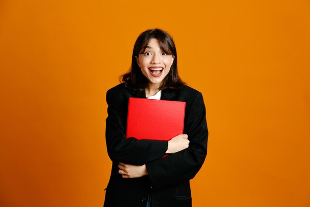 Surprised holding folder young beautiful female wearing black jacket isolated on orange background