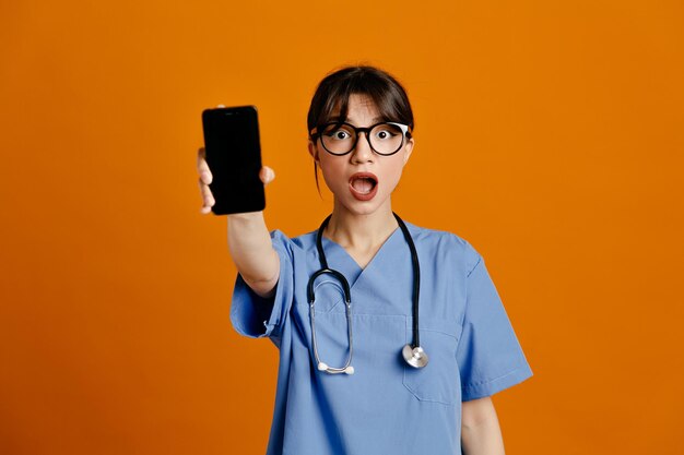 Surprised holding out phone at camera young female doctor wearing uniform fith stethoscope isolated on orange background