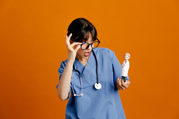 オレンジ色の背景に分離された制服の聴診器を身に着けているおもちゃの若い女性医師を保持している驚いた