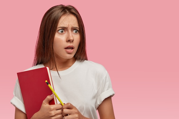 Foto gratuita giovane donna sorpresa e inorridita con i capelli lisci e scuri, tiene in mano un libro di testo rosso, una matita, si sente perplessa di ricevere molti compiti