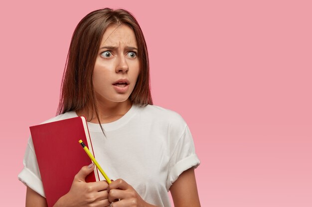 Surprised horrified young woman with dark straight hair, holds red textbook, pencil, feels puzzled of recieving many tasks