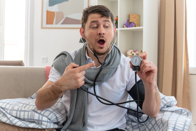 surprised ill man with scarf around neck measuring his pressure with and pointing at sphygmomanometer sitting on couch at living room