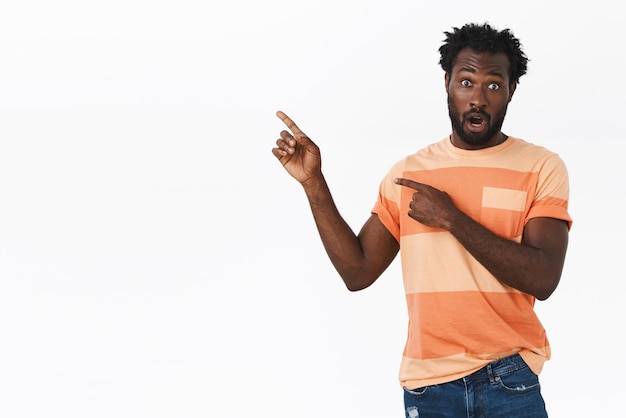 Surprised and impressed african american bearded guy in striped t-shirt, open mouth fascinated, gasping shocked as pointing upper left corner
