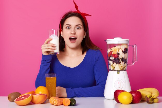 Surprised impressed brunette female sits at table with opened mouth widely, holding glass of milkshake in right hand, cooking other fruit mixture in white blender. Healthy lifestyle concept.