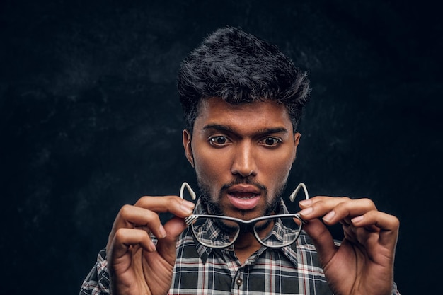 Surprised Indian guy lowers his glasses and realizes that now he can see without glasses. Studio photo against a dark textured wall