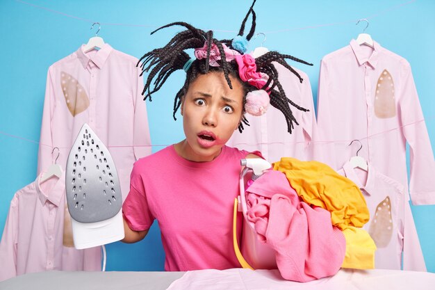 Surprised indignant dark skinned woman has funny hairstyle poses with stack of laundry going to iron does domestic work poses near ironing board against blue wall