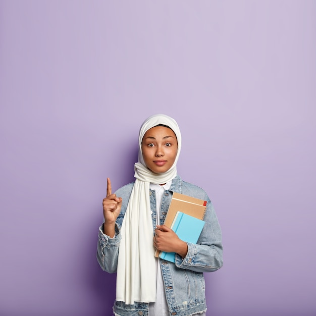 Free photo surprised intriguing woman in headwear points upwards and looks with interest, shows blank space above for your advertisement or information, carries diary and spiral notebook. muslim religion