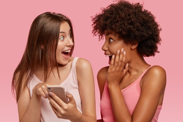 Surprised joyful interracial friends look happily at screen of cell phone, read something unbelievable, pose together against pink wall, make shopping online. Ethnicity and technology.