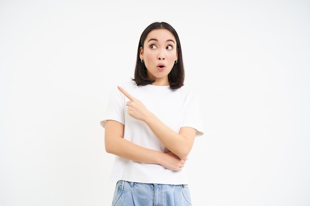 Free photo surprised korean girl points left and looks with amazed face expression stands in tshirt isolated on