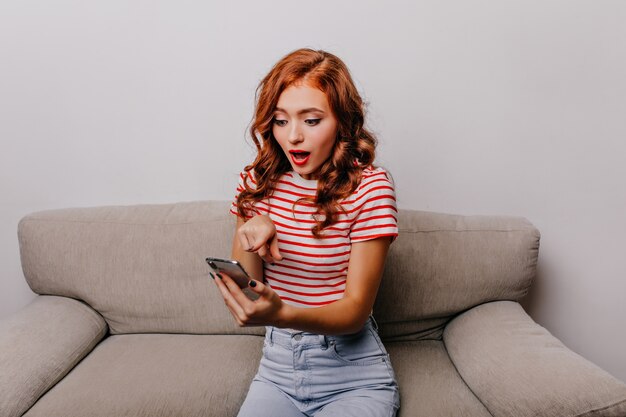 Surprised lady looking at phone screen while sitting on couch. Ginger girl in striped t-shirt posing with smartphone.
