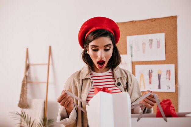 Surprised lady in red beret looks into shopping bag. Young woman with dark hair with bright lipstick in striped stylish clothes posing on camera.