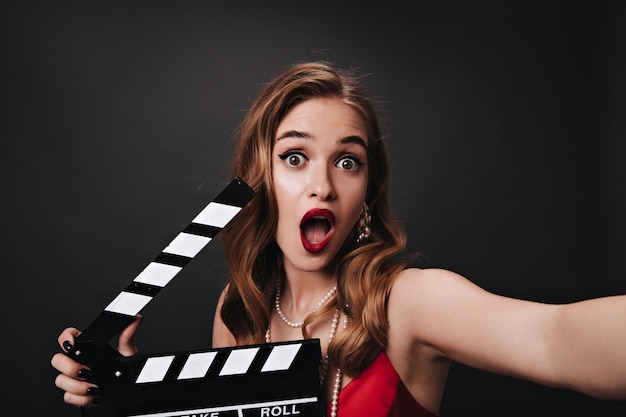 Free photo surprised lady with beautiful makeup holding clapperboard and taking selfie shocked actress in red silk top posing on black background