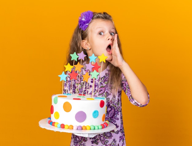 surprised little blonde girl holding birthday cake and keeping hand close to mouth calling someone isolated on orange wall with copy space