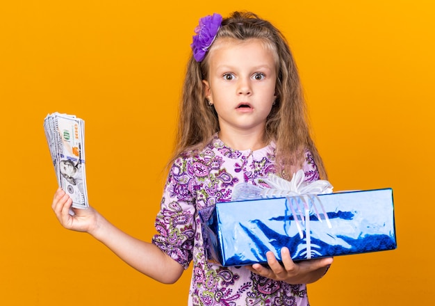 surprised little blonde girl holding gift box and money isolated on orange wall with copy space