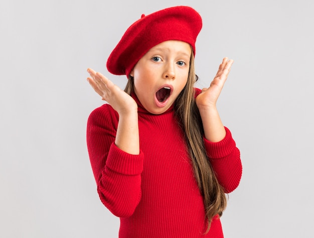 Piccola ragazza bionda sorpresa che indossa berretto rosso che tiene le mani vuote in su guardando la parte anteriore con la bocca aperta isolata sul muro bianco con spazio di copia