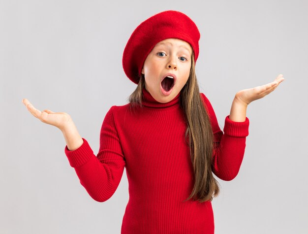 Surprised little blonde girl wearing red beret showing empty hands in air looking at camera isolated on white wall with copy space